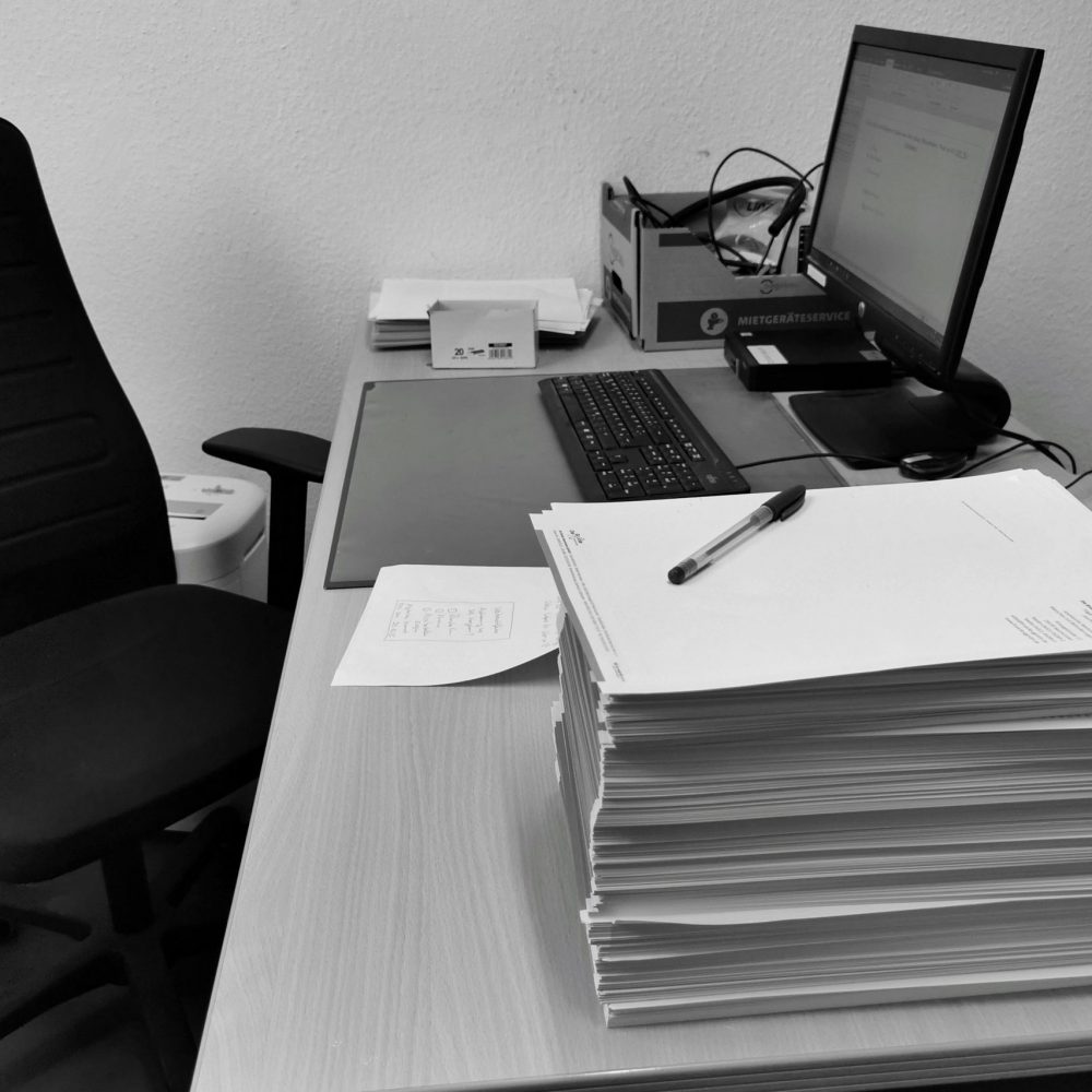 Black and white image of a cluttered office desk with a computer, chair, and paperwork.